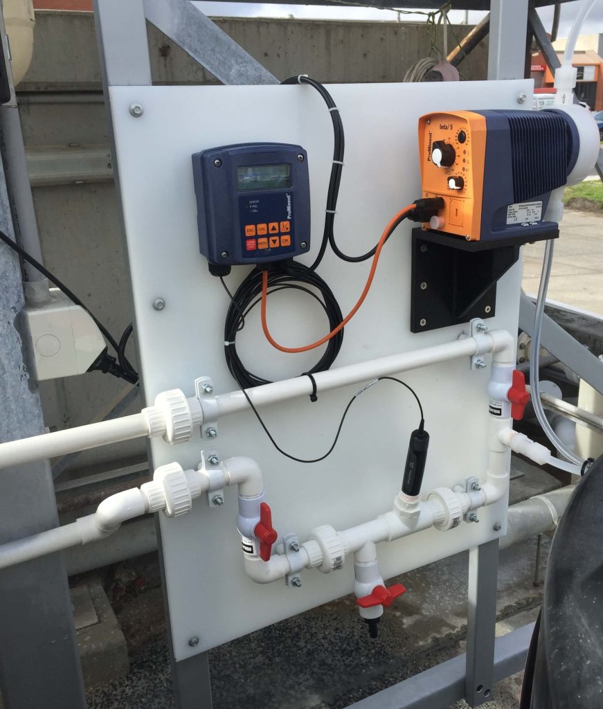 Wall-mounted wastewater monitoring panel with inline sensor, control display, and automatic chemical dosing pump connected to pipework and isolation valves for water quality control.