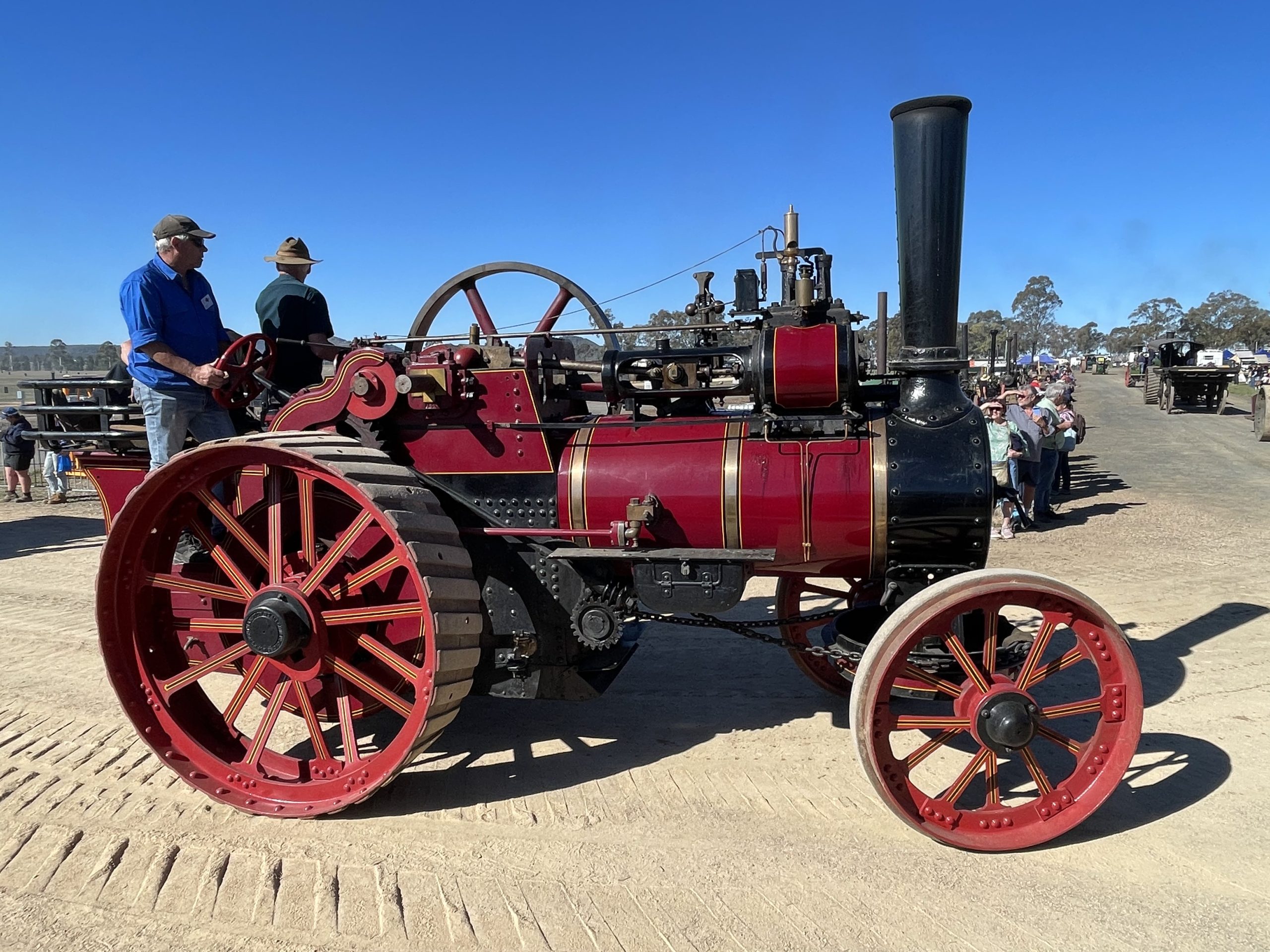 Red and black steam traction engine with gold trim driving along a dirt track at a heritage machinery rally, with operators standing on the platform and spectators watching in the background.