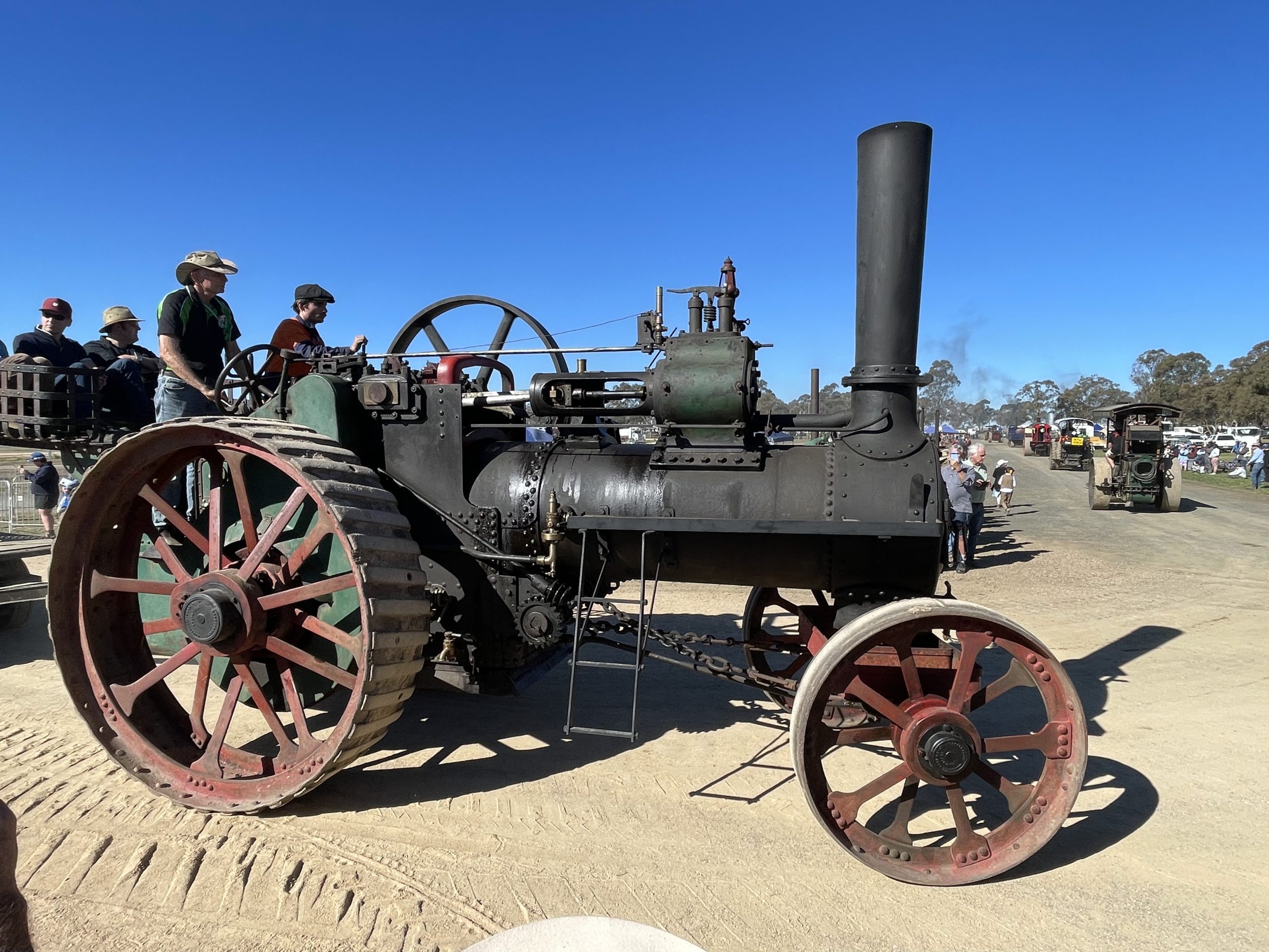 Steam traction engine driving along a dirt track during a heritage machinery rally, with spectators watching under a clear blue sky.