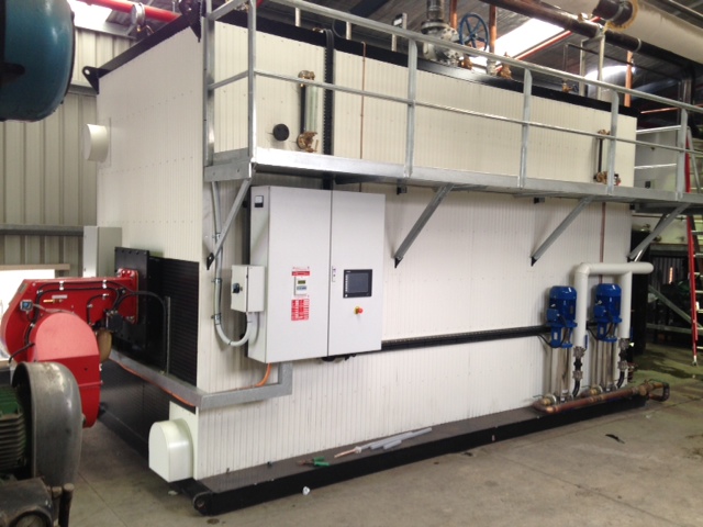 Large industrial boiler installed on a customer site, featuring a red burner assembly, control panel, circulation pumps, and overhead pipework with an access platform above the unit.